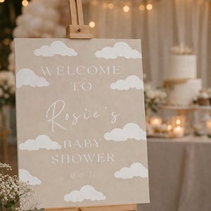 Op de afbeelding: Een beige bord op een houten ezel met witte wolkuitsparingen. Op het bord staat "WELCOME TO Rosie's BABY SHOWER 18-01-20". De achtergrond omvat een taart, bloemen en lichtslingers.