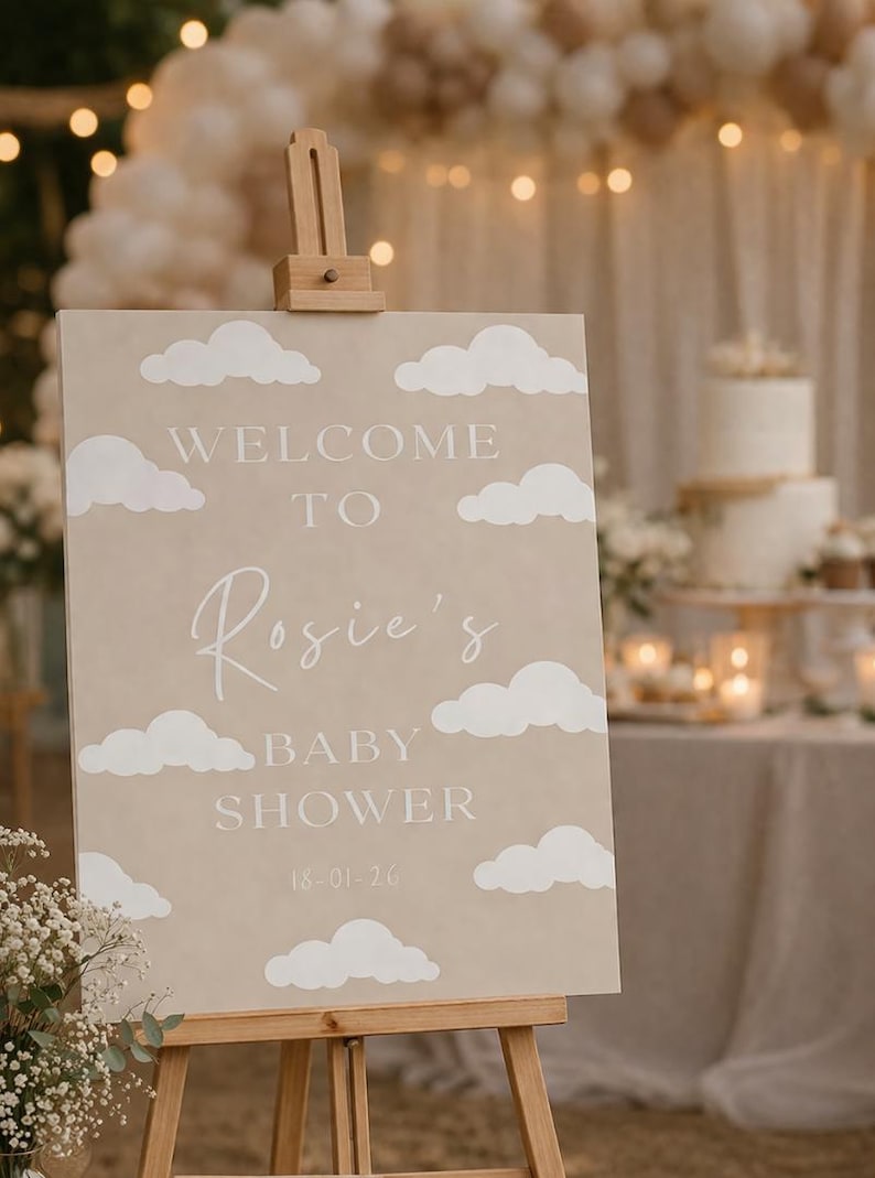 Op de afbeelding: Een beige bord op een houten ezel met witte wolkuitsparingen. Op het bord staat "WELCOME TO Rosie's BABY SHOWER 18-01-20". De achtergrond omvat een taart, bloemen en lichtslingers.