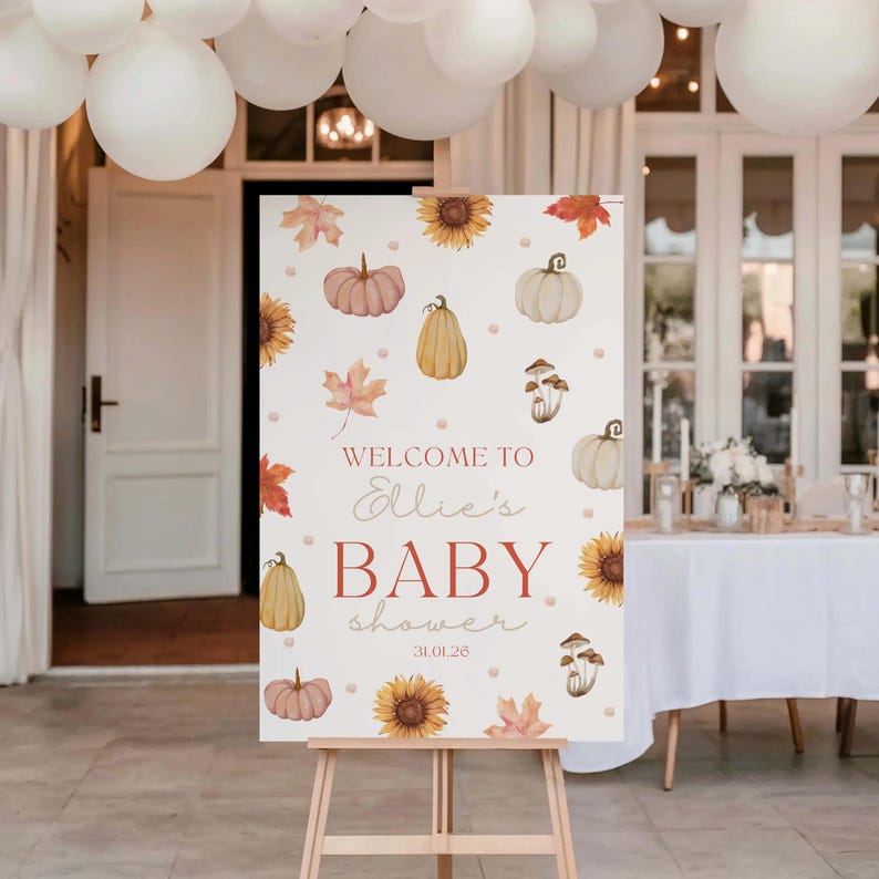 May include: A welcome sign for a baby shower, featuring a fall theme with pumpkins, sunflowers, and leaves. The sign reads "Welcome to Ellie's BABY shower 31.01.26" in a decorative font, displayed on a wooden easel.