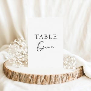 K&ouml;nnte beinhalten: Eine wei&szlig;e, rechteckige Tischnummernkarte mit dem schwarzen Aufdruck TABLE One in einer Kombination aus Serifenschrift und Schreibschrift. Die Karte steht auf einer runden Holzscheibe mit nat&uuml;rlicher Rinde und kleinen wei&szlig;en Bl&uuml;ten.