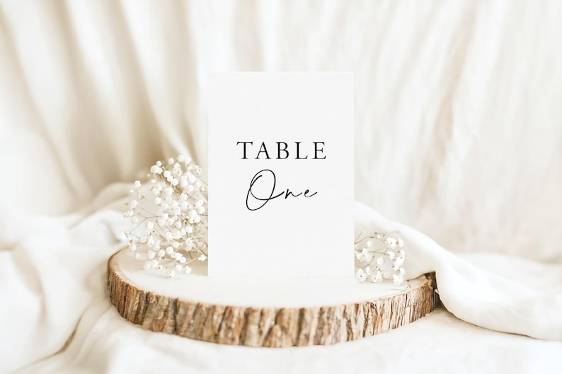 K&ouml;nnte beinhalten: Eine wei&szlig;e, rechteckige Tischnummernkarte mit dem schwarzen Aufdruck TABLE One in einer Kombination aus Serifenschrift und Schreibschrift. Die Karte steht auf einer runden Holzscheibe mit nat&uuml;rlicher Rinde und kleinen wei&szlig;en Bl&uuml;ten.