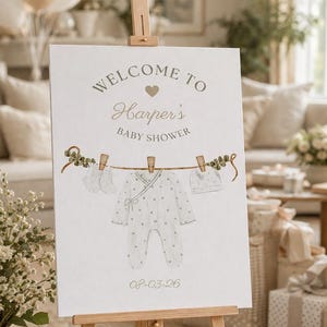 May include: A baby shower sign on a wooden easel. The sign reads "Welcome to Harper's Baby Shower" with a heart graphic. A clothesline illustration displays baby clothes, including a onesie and hat. The date "08-03-26" is printed at the bottom.