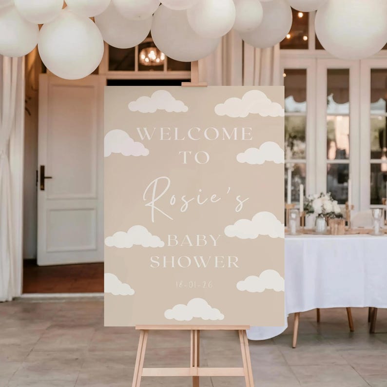 Op de afbeelding: Een beige welkomstbord voor een babyshower, met witte wolkenillustraties en de tekst "Welcome to Rosie's Baby Shower 16-01-26". Het bord staat op een houten ezel, met witte ballonnen erboven.