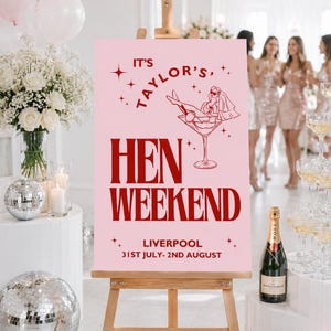 May include: A pink sign on a wooden easel advertises a hen weekend in Liverpool. The sign has red text and a martini glass illustration. Decorations include balloons, flowers, disco balls, and champagne.