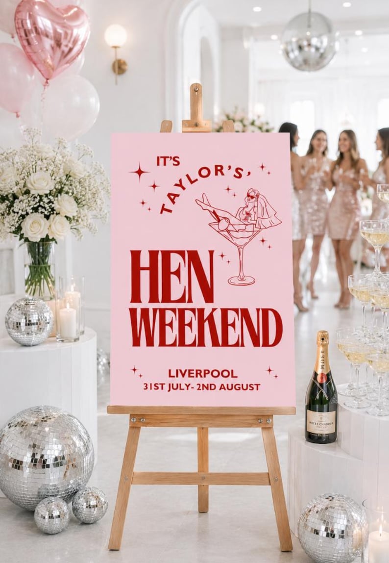May include: A pink sign on a wooden easel advertises a hen weekend in Liverpool. The sign has red text and a martini glass illustration. Decorations include balloons, flowers, disco balls, and champagne.