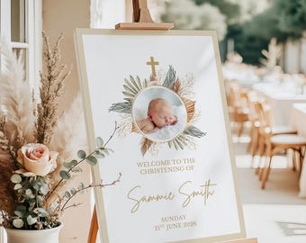 May include: A rectangular christening welcome sign on a wooden easel featuring a circular photo of a baby, a gold cross, and dried botanical illustrations. The text reads: Welcome to the christening of Sammie Smith, Sunday 21st June 2026.