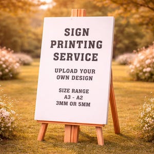May include: A white sign on a wooden easel, displaying the text "SIGN PRINTING SERVICE". The sign also reads "UPLOAD YOUR OWN DESIGN" and "SIZE RANGE A3 - A2 3MM OR 5MM". The easel is set on a grassy area with a blurred background.