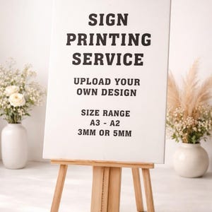 May include: A wooden easel displays a white sign with black text: "SIGN PRINTING SERVICE, UPLOAD YOUR OWN DESIGN, SIZE RANGE A3 - A2, 3MM OR 5MM." The easel is in front of a white background with floral arrangements.