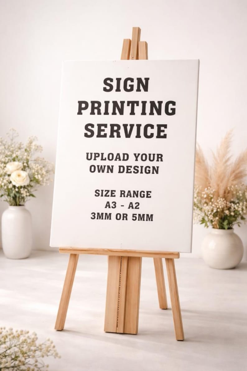 May include: A wooden easel displays a white sign with black text: "SIGN PRINTING SERVICE, UPLOAD YOUR OWN DESIGN, SIZE RANGE A3 - A2, 3MM OR 5MM." The easel is in front of a white background with floral arrangements.