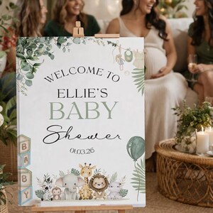 Pu&ograve; includere: Un cavalletto in legno espone un cartello di benvenuto per un baby shower. Il cartello recita "Welcome to Ellie's Baby Shower" con un design botanico e illustrazioni di animali per bambini. Sono presenti anche un palloncino verde e la data 01.03.26.