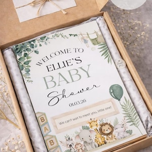 Pu&ograve; includere: Cartello per baby shower formato A4 in una scatola. Il cartello recita "Benvenuta al Baby Shower di Ellie" con la data 01.03.26. Il cartello ha una grafica a palloncino verde e un bordo di animali della giungla. La scatola include un biglietto "Grazie".