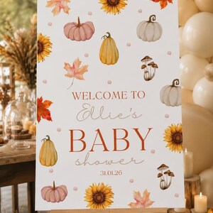 May include: A white welcome sign on an easel, decorated with watercolor pumpkins, sunflowers, and fall leaves. The sign reads "Welcome to Ellie's Baby Shower 31.01.26" in a mix of fonts and colors, perfect for a fall-themed baby shower.