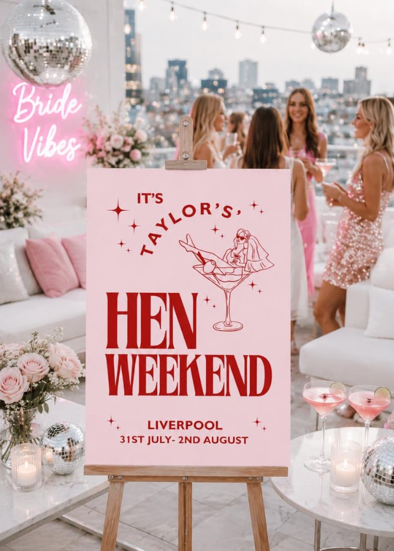 May include: A pink sign on an easel with the text "It's Taylor's Hen Weekend Liverpool 31st July- 2nd August". The sign has a red illustration of a bride in a martini glass. The background shows a rooftop party with women.