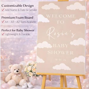 Op de afbeelding: Een gepersonaliseerd welkomstbord voor een babyshower op een houten ezel. Het beige bord heeft witte wolkenaccenten en de tekst "Welcome to Rosie's Baby Shower" met de datum. Volledig aanpasbaar.