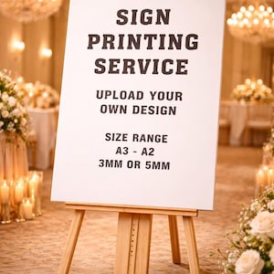 May include: A wooden easel displays a white sign with black text: "SIGN PRINTING SERVICE, UPLOAD YOUR OWN DESIGN, SIZE RANGE A3 - A2, 3MM OR 5MM." The sign is in a well-lit room with chandeliers and floral arrangements.
