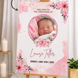 May include: A christening sign on a wooden easel, featuring a baby's photo within a pink floral frame. The sign reads "Welcome to the Christening of Louise Teller" and the date "Sunday | June 14th | 2026".