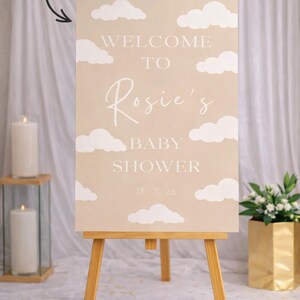 Op de afbeelding: Een welkomstbord voor een babyshower op een houten ezel. Het beige bord toont witte wolkenillustraties en de tekst "Welcome to Rosie's Baby Shower 18.01.26". Kaarsen en bloemen sieren de achtergrond.