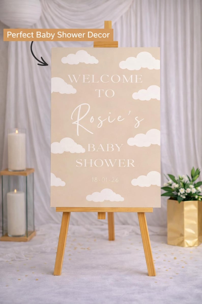 Op de afbeelding: Een welkomstbord voor een babyshower op een houten ezel. Het beige bord toont witte wolkenillustraties en de tekst "Welcome to Rosie's Baby Shower 18.01.26". Kaarsen en bloemen sieren de achtergrond.