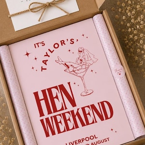 May include: A hen weekend gift box with a pink and red design. The box contains a pink poster with the text "IT'S TAYLOR'S, HEN WEEKEND" and a drawing of a woman in a martini glass. The box also includes a card that says "Thank you".