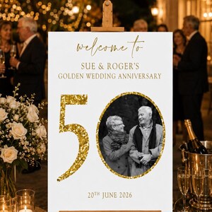 May include: A white sign on a wooden easel with gold text that reads "welcome to SUE & ROGER'S GOLDEN WEDDING ANNIVERSARY". A gold glitter "50" and a black and white photo of a couple are also on the sign. The date "20th JUNE 2026" is at the bottom.