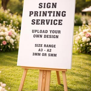 May include: A white sign on a wooden easel, displaying the text "SIGN PRINTING SERVICE." The sign also includes "UPLOAD YOUR OWN DESIGN" and "SIZE RANGE A3 - A2 3MM OR 5MM." The easel is on a grassy lawn with flowers in the background.