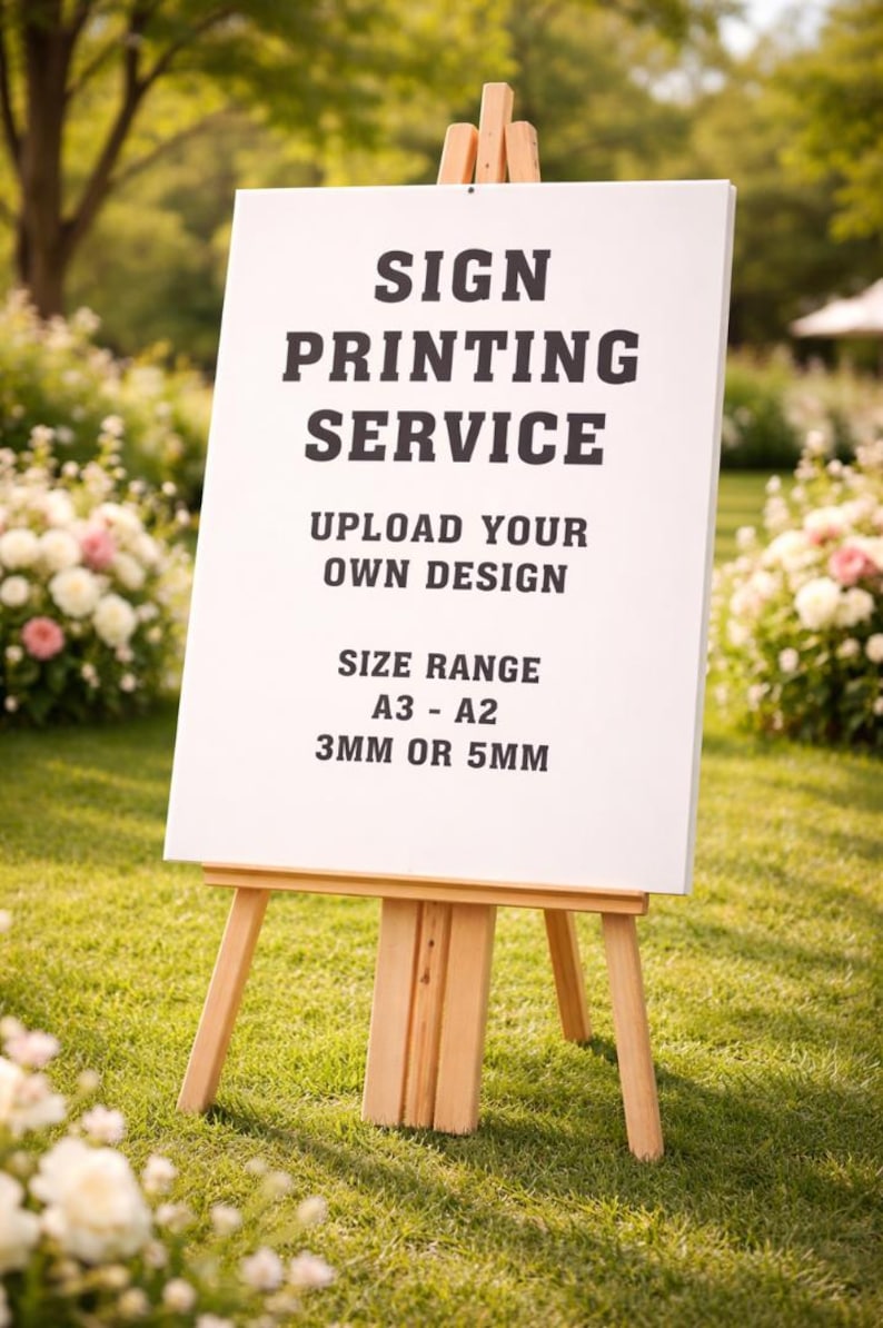 May include: A white sign on a wooden easel, displaying the text "SIGN PRINTING SERVICE." The sign also includes "UPLOAD YOUR OWN DESIGN" and "SIZE RANGE A3 - A2 3MM OR 5MM." The easel is on a grassy lawn with flowers in the background.