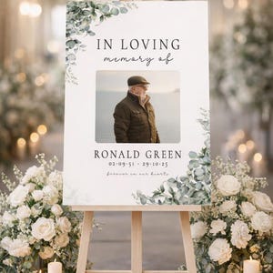 Peut inclure: Un panneau comm&eacute;moratif sur un chevalet en bois, avec le texte "IN LOVING memory of" au-dessus d'une photo d'un homme. Le panneau comprend le nom "RONALD GREEN" et des dates, avec des motifs floraux. Des fleurs blanches et des bougies entourent le chevalet.