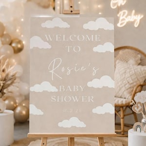Op de afbeelding: Een beige bord op een houten ezel met witte wolken. Op het bord staat "WELCOME TO Rosie's BABY SHOWER" met de datum 16-01-26. De achtergrond toont ballonnen en een neon "Oh Baby" bord.