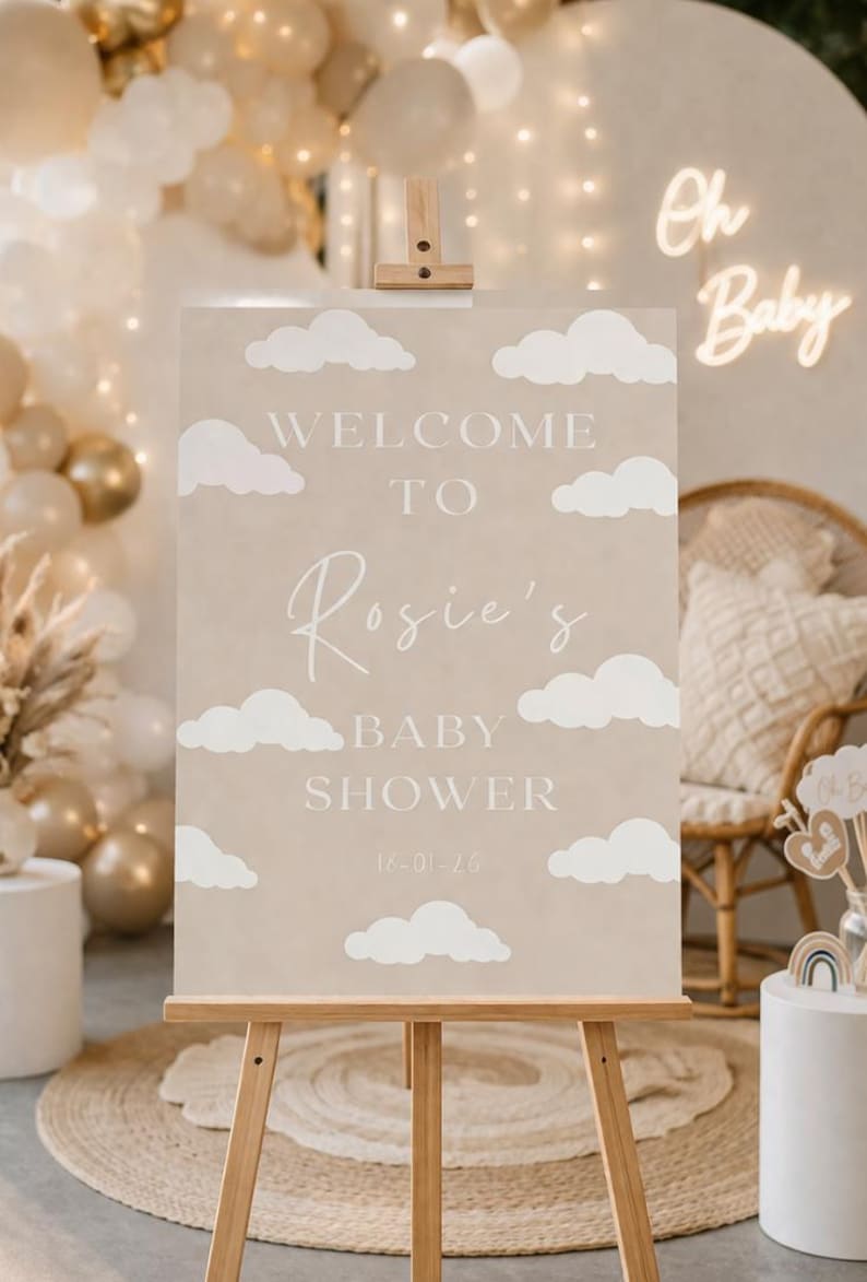 Op de afbeelding: Een beige bord op een houten ezel met witte wolken. Op het bord staat "WELCOME TO Rosie's BABY SHOWER" met de datum 16-01-26. De achtergrond toont ballonnen en een neon "Oh Baby" bord.
