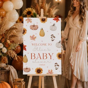 May include: A wooden easel displays a sign with a fall-themed baby shower design. The sign features watercolor pumpkins, sunflowers, and leaves. Text reads "Welcome to Ellie's BABY shower 31.01.26". Balloons and floral arrangements decorate the background.