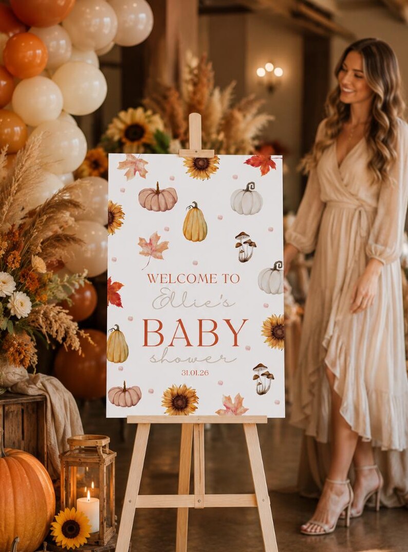 May include: A wooden easel displays a sign with a fall-themed baby shower design. The sign features watercolor pumpkins, sunflowers, and leaves. Text reads "Welcome to Ellie's BABY shower 31.01.26". Balloons and floral arrangements decorate the background.