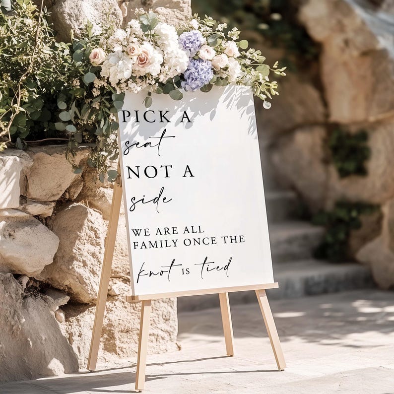 K&ouml;nnte beinhalten: Ein wei&szlig;es, rechteckiges Hochzeitsschild auf einer hellen Holzstaffelei mit schwarzem Text: PICK A seat NOT A side WE ARE ALL FAMILY ONCE THE knot is tied. Das Schild ist mit einem Blumenarrangement aus wei&szlig;en und blauen Bl&uuml;ten verziert.