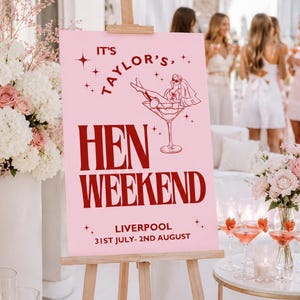 May include: A pink sign on a wooden easel announces a "Hen Weekend" in Liverpool. The sign features a graphic of a woman in a martini glass and the text "It's Taylor's". The event dates are 31st July to 2nd August.
