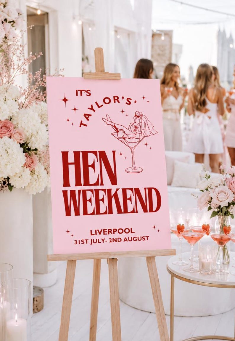 May include: A pink sign on a wooden easel announces a "Hen Weekend" in Liverpool. The sign features a graphic of a woman in a martini glass and the text "It's Taylor's". The event dates are 31st July to 2nd August.