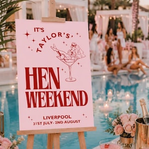 May include: A pink sign on a wooden easel reads "It's Taylor's, Hen Weekend, Liverpool, 31st July-2nd August". The sign features a martini glass with a bride illustration. The scene is set by a pool with cocktails and flowers.