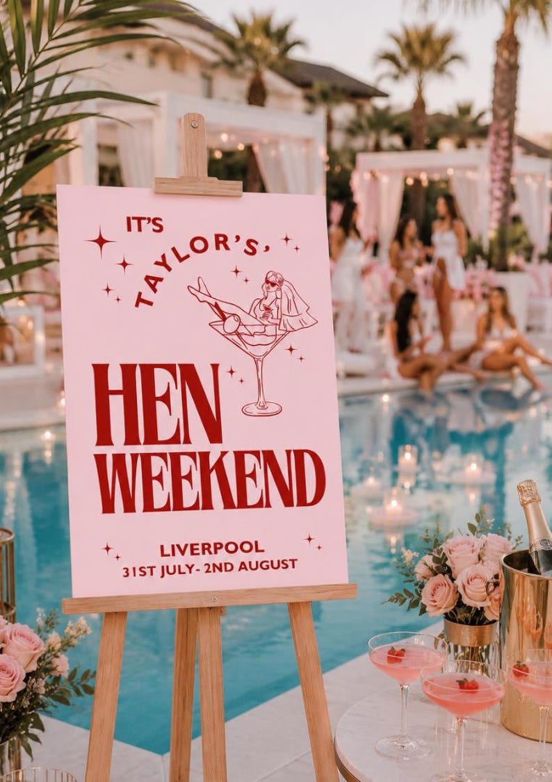 May include: A pink sign on a wooden easel reads "It's Taylor's, Hen Weekend, Liverpool, 31st July-2nd August". The sign features a martini glass with a bride illustration. The scene is set by a pool with cocktails and flowers.