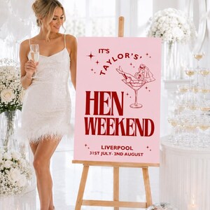May include: A pink sign on a wooden easel with the text "IT'S TAYLOR'S HEN WEEKEND LIVERPOOL 31ST JULY-2ND AUGUST". The sign has a graphic of a woman in a martini glass. A woman in a white dress is holding a champagne glass.