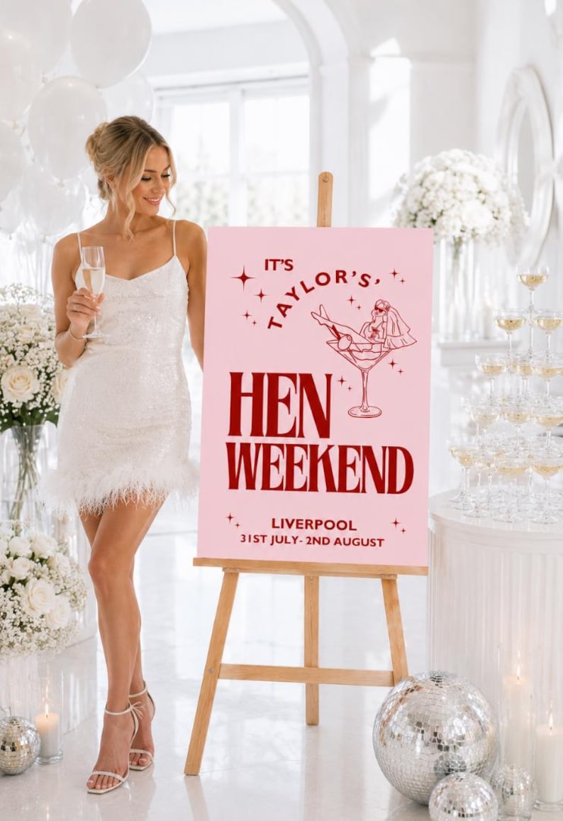 May include: A pink sign on a wooden easel with the text "IT'S TAYLOR'S HEN WEEKEND LIVERPOOL 31ST JULY-2ND AUGUST". The sign has a graphic of a woman in a martini glass. A woman in a white dress is holding a champagne glass.