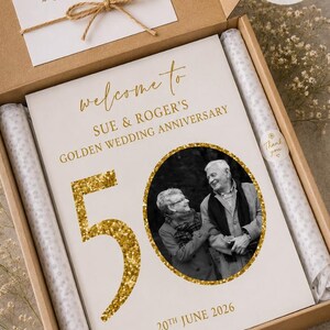 May include: A personalized golden wedding anniversary gift box. The box contains a card with the text "welcome to SUE & ROGER'S GOLDEN WEDDING ANNIVERSARY" and a photo of the couple. The number "50" is in gold glitter. The date "20th June 2026" is also included.