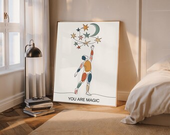 May include: A framed art print featuring a minimalist line drawing of a person reaching for stars and a crescent moon. The artwork is in a gold frame and includes the text "YOU ARE MAGIC" at the bottom. The color palette includes beige, orange, blue, and green.