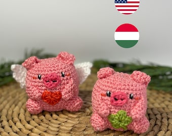 Könnte beinhalten: Zwei handgefertigte rosa gehäkelte Schweinchenfiguren. Ein Schwein hat weiße Flügel und ein rotes Herz, das andere hält ein grünes vierblättriges Kleeblatt. Die Schweine haben kleine schwarze Augen und rosa Schnauzen. Der Hintergrund ist ein geflochtener Korb.