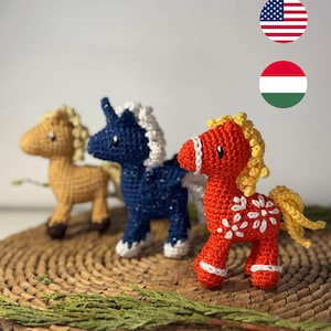 May include: Three handmade crochet horse figurines in tan, navy blue, and red. Each horse has a white mane and tail. The red horse has white floral accents. The horses are displayed on a woven mat with greenery.