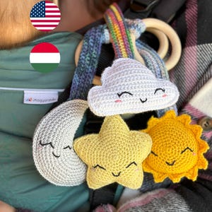 May include: A crochet mobile featuring a white cloud, a yellow star, a golden sun, and a grey crescent moon, all with embroidered smiling faces. The figures are attached to a rainbow-coloured strap and a smooth, round wooden ring for hanging.