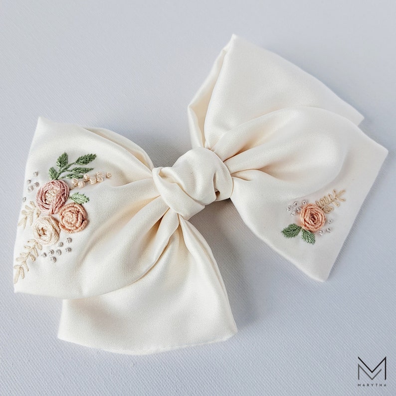 May include: A white satin bow with embroidered floral details in shades of pink and green. The bow is tied in a classic shape with a center knot.