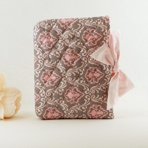 May include: A rectangular quilted fabric cover featuring a brown and pink damask pattern with a diamond-stitched texture. The cover is accented with a light pink satin ribbon tied into a bow on the right side, providing a soft contrast to the fabric.