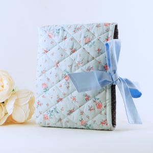 May include: A quilted fabric book cover featuring a light blue background with a small pink floral pattern. The cover is secured with a solid light blue ribbon tied in a bow on the side, revealing a dark brown interior lining with white dots.
