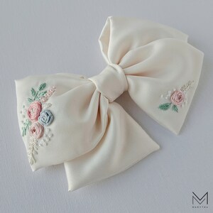 May include: A white satin bow with delicate floral embroidery in shades of pink, blue, and green. The bow is perfect for adding a touch of elegance to any occasion.