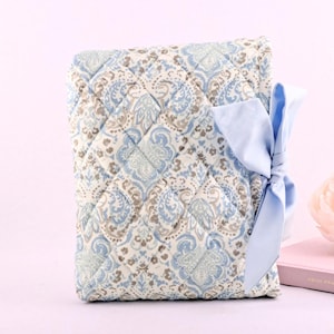 May include: A rectangular quilted fabric cover featuring a light blue, beige, and white paisley pattern. The cover is secured with a light blue satin ribbon tied into a bow on the right side. The quilted texture consists of a diamond-stitched grid.
