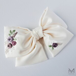 May include: A large, ivory satin bow with delicate floral embroidery in shades of purple and green. The bow is tied in a classic shape with a center knot.