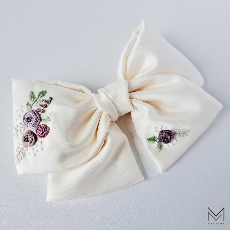 May include: A large, ivory satin bow with delicate floral embroidery in shades of purple and green. The bow is tied in a classic shape with a center knot.